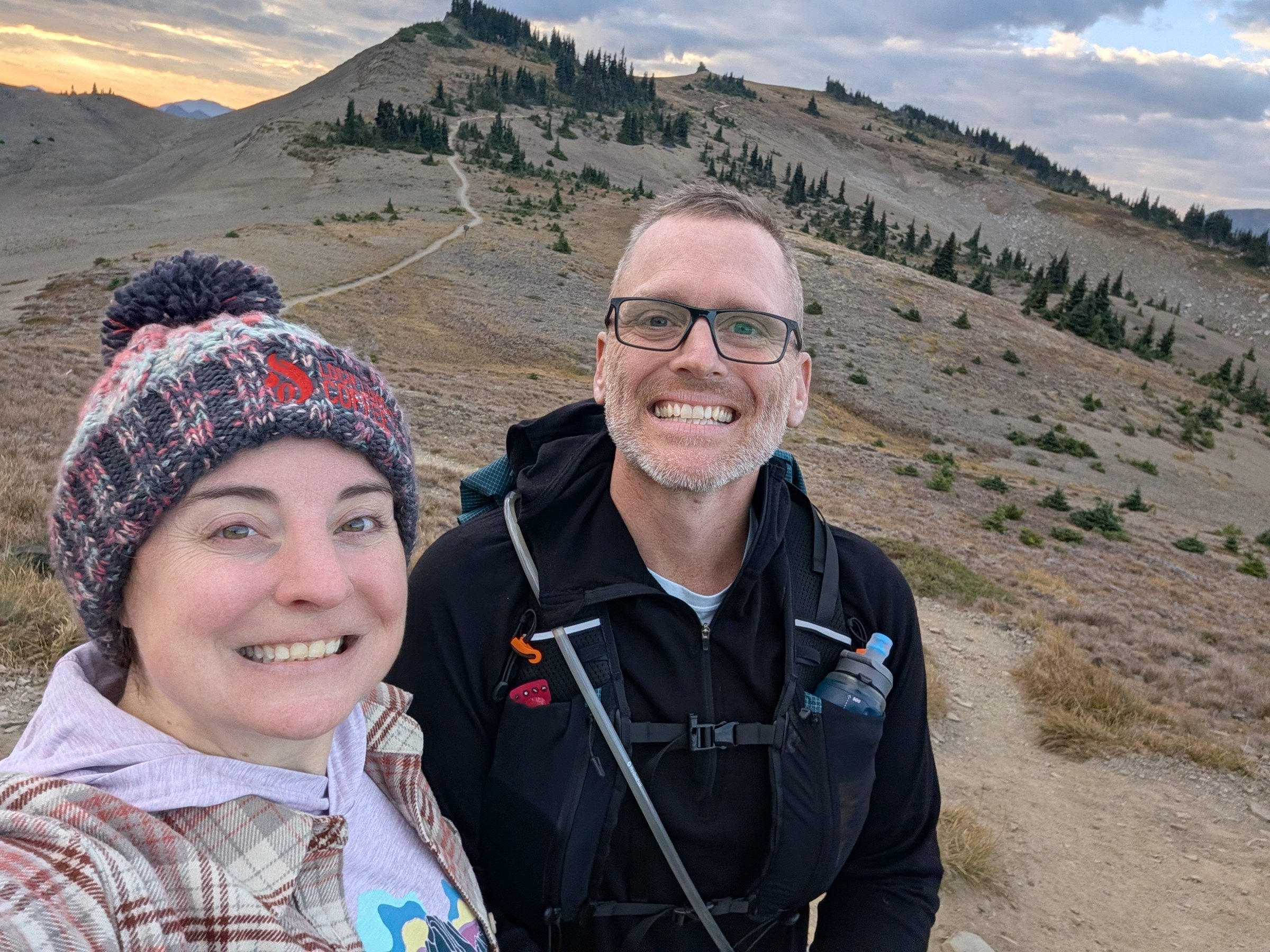 Adam and Jeney smiling in a selfie at Obstruction Point at sunrise, with the trail climbing a ridge behind them