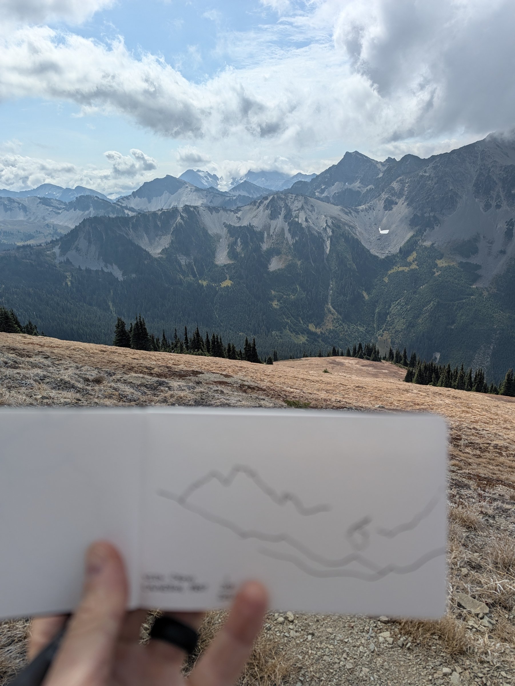 Adam holding a small sketchbook with a pen drawing of the ridgeline up against the real mountains behind it